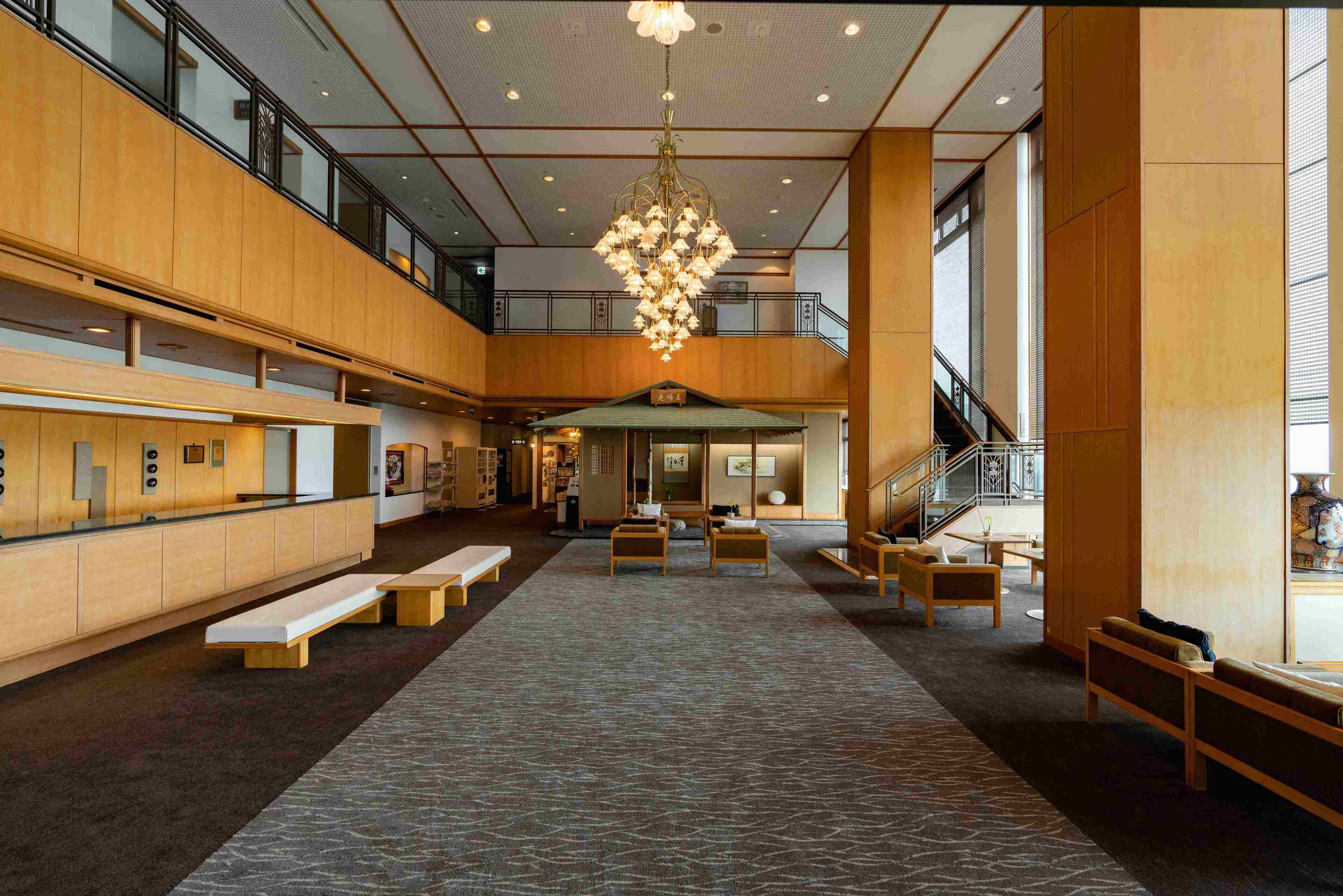 A spacious lobby with a high ceiling, a central chandelier, wooden benches, and a staircase leading to an upper level.