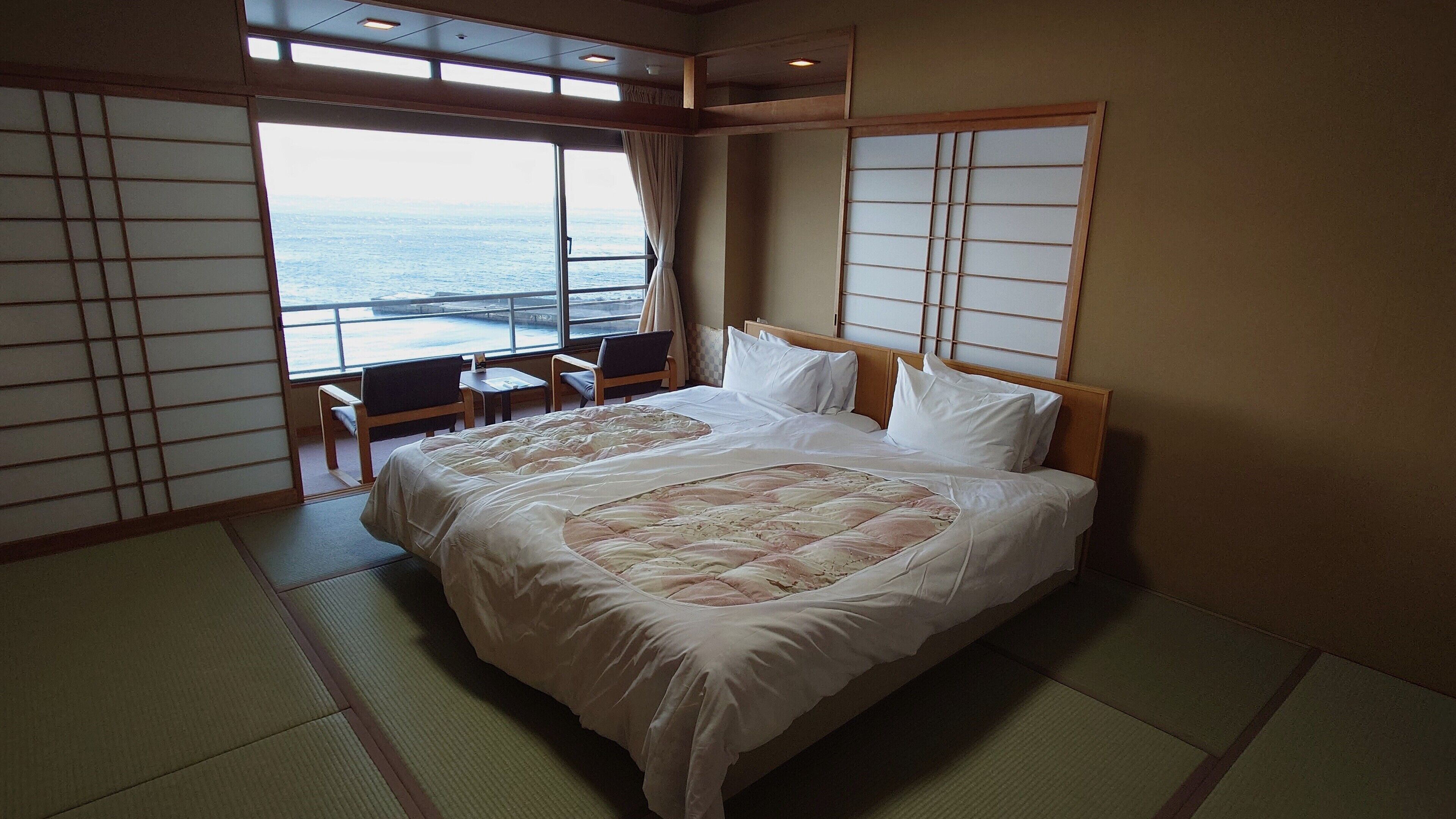 Habitación de hotel con una cama grande, dos mesitas de noche, vista al mar a través de una ventana y puertas correderas tradicionales japonesas.