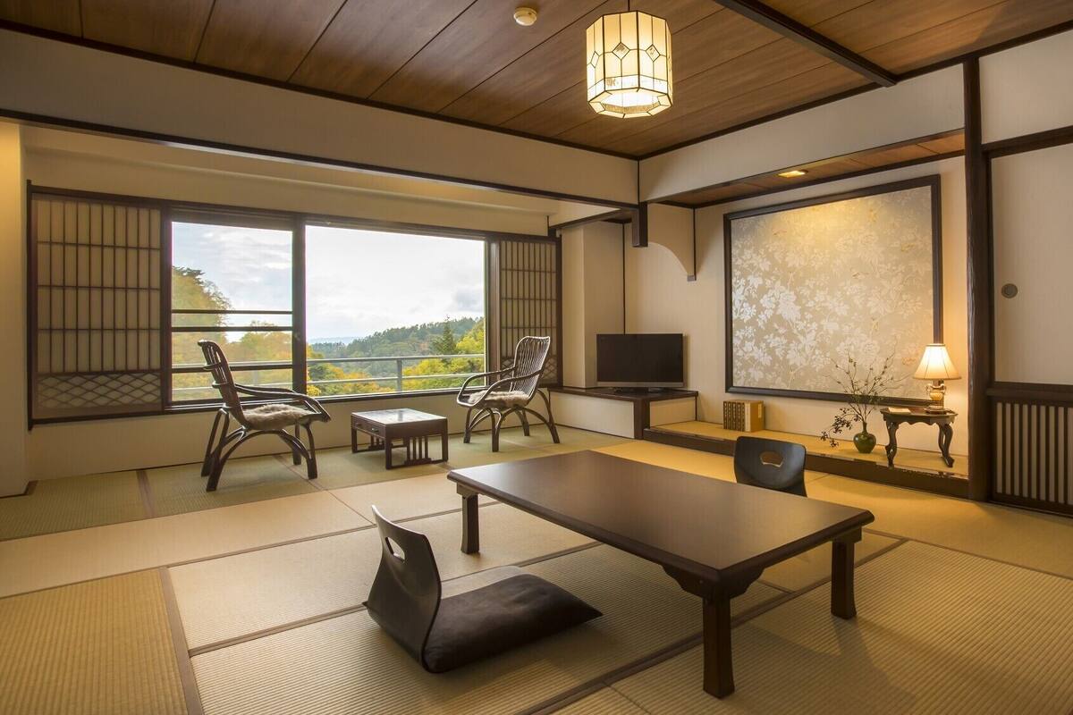 Quarto japonês tradicional com tatames, uma mesa baixa, cadeiras, televisão e vista para o exterior.