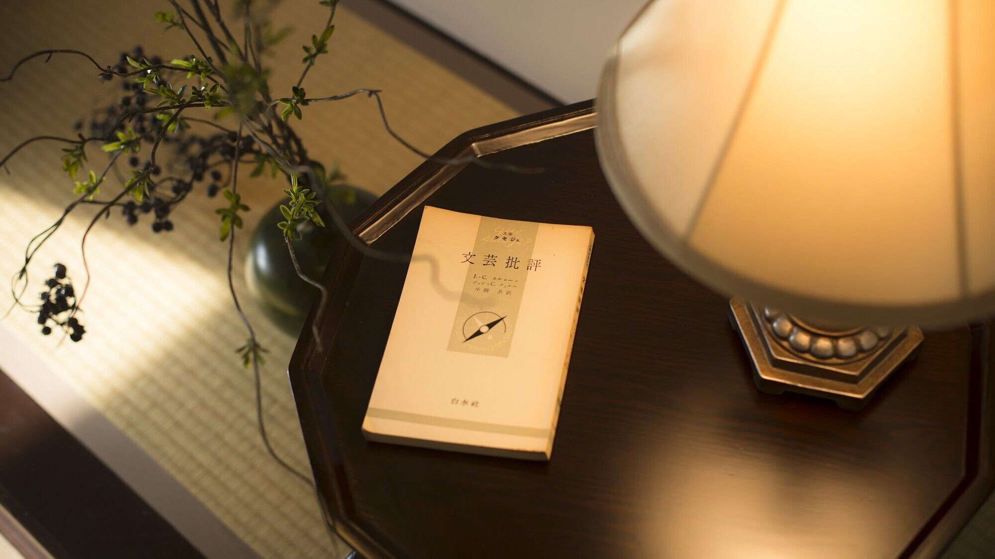 Un llibre amb el títol '文芸批評' sobre una taula auxiliar de fusta fosca amb una làmpada.