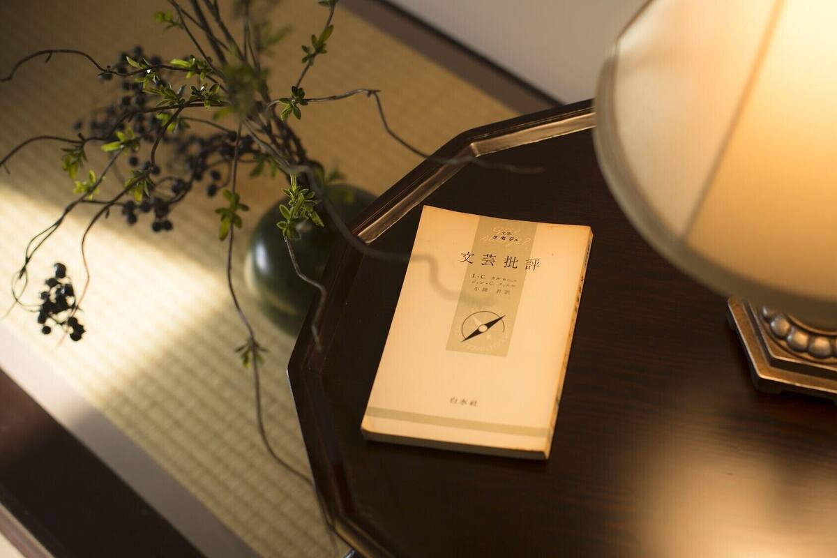 Um livro intitulado '文芸批評' sobre uma mesa de cabeceira de madeira escura com uma lâmpada.
