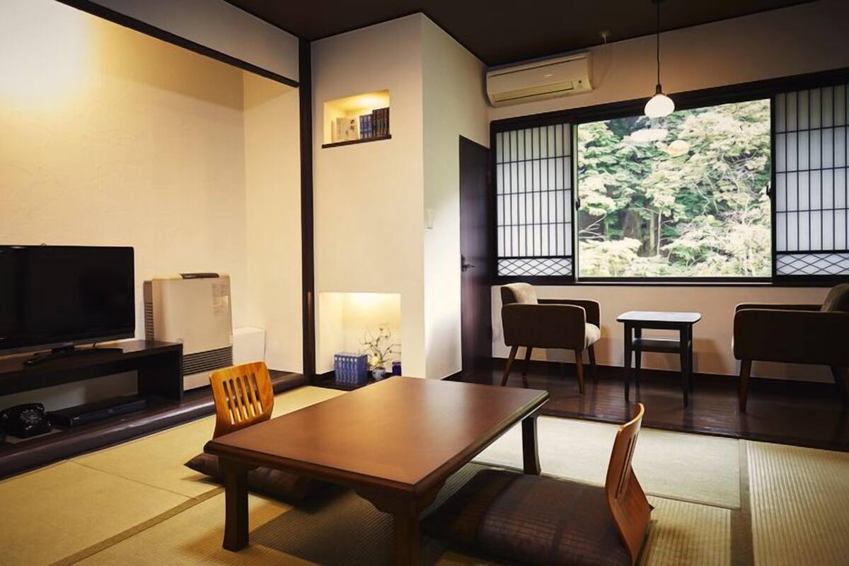 Quarto japonês tradicional com tatames, televisão, aquecedor e vista para árvores através de um painel shoji.