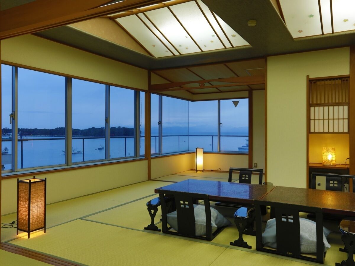 A room with tatami flooring, a low table, chairs, a large window with a view of water, and a traditional Japanese lantern.