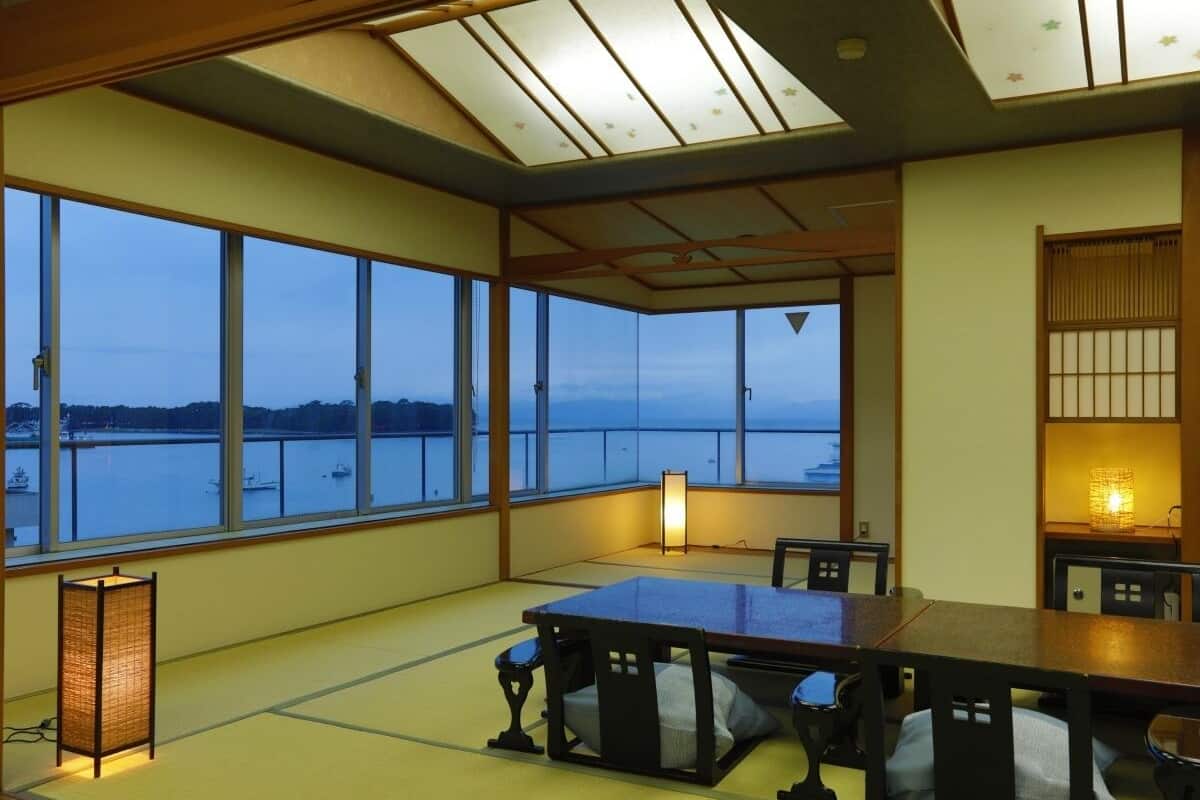 Una habitación con suelo de tatami, una mesa baja, sillas, un ventanal con vistas al agua y un farolillo japonés tradicional.
