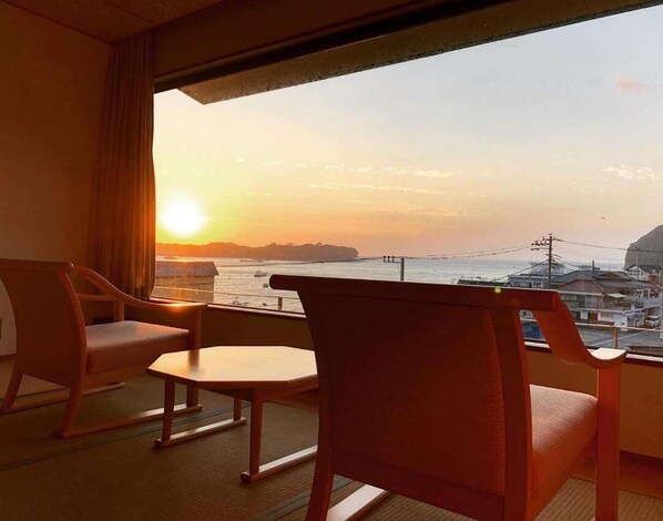 A room with a view of the sunset over the sea, featuring two wooden chairs and a small table.
