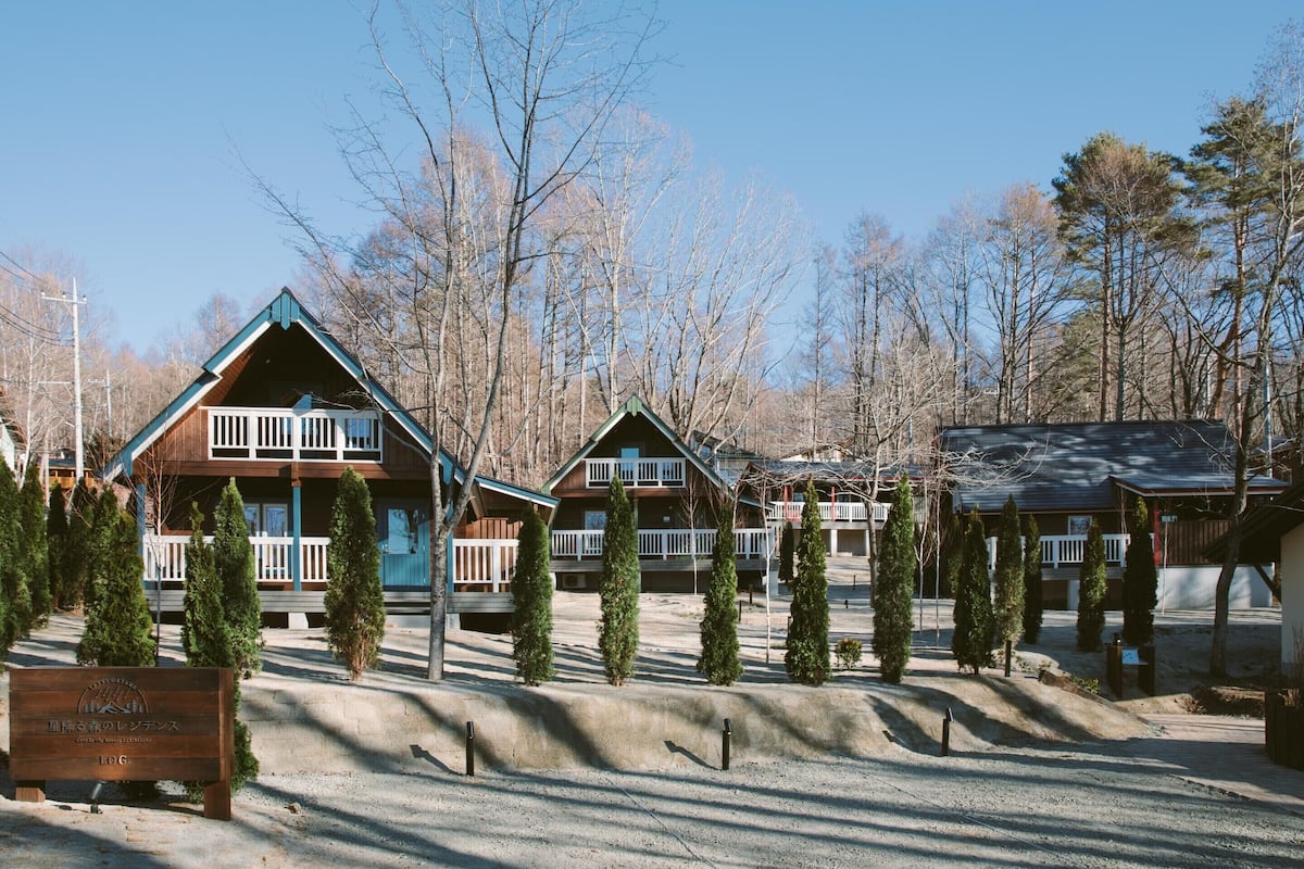 A row of log cabin-style cabins with balconies, surrounded by trees and a gravel path.
