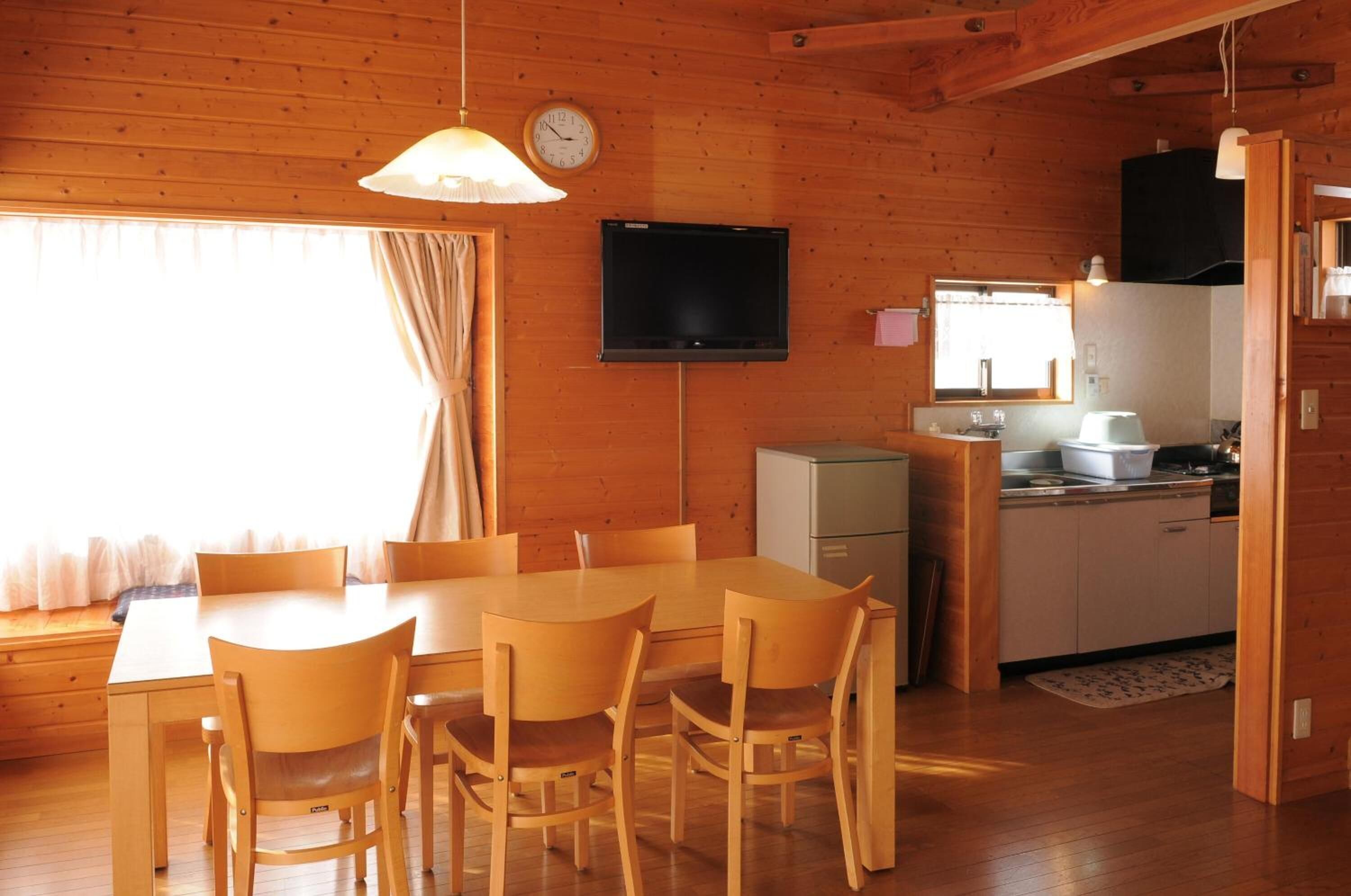 Interno di una baita in legno con tavolo da pranzo, sedie, televisore, angolo cottura e finestra con tende.