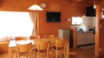 Interno di una baita in legno con tavolo da pranzo, sedie, televisore, angolo cottura e finestra con tende.