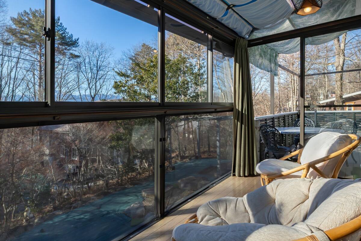 A glass-enclosed room with a view of a forest and a body of water. There is a wicker chair and a table with chairs.