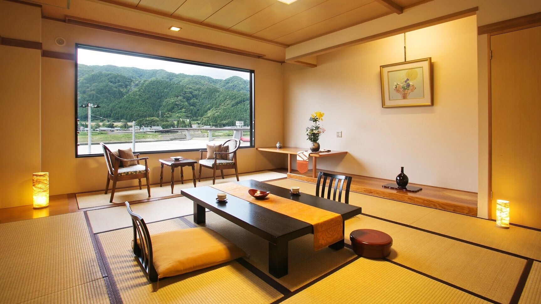 A traditional Japanese room with tatami flooring, a low table, chairs, a view of mountains, and a framed picture on the wall.