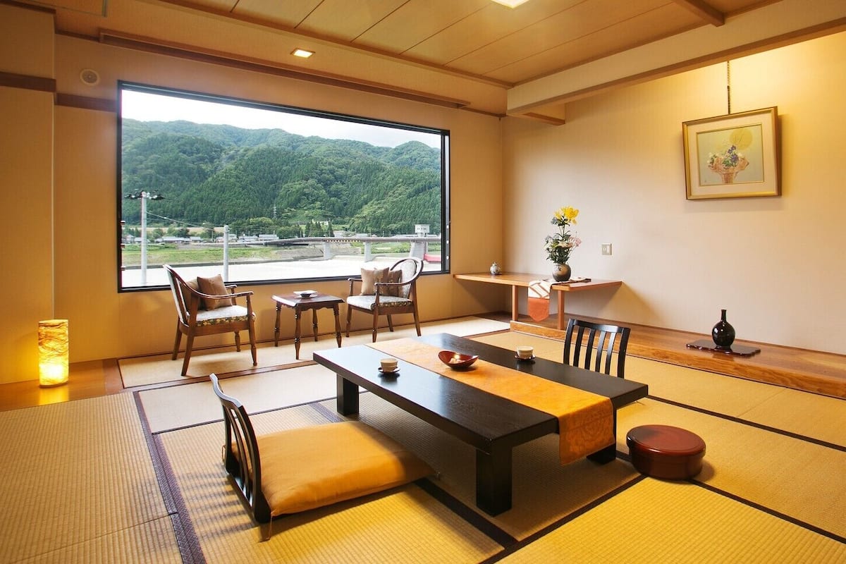 Una habitación japonesa tradicional con suelo de tatami, una mesita baja, sillas, vista a las montañas y un cuadro en la pared.