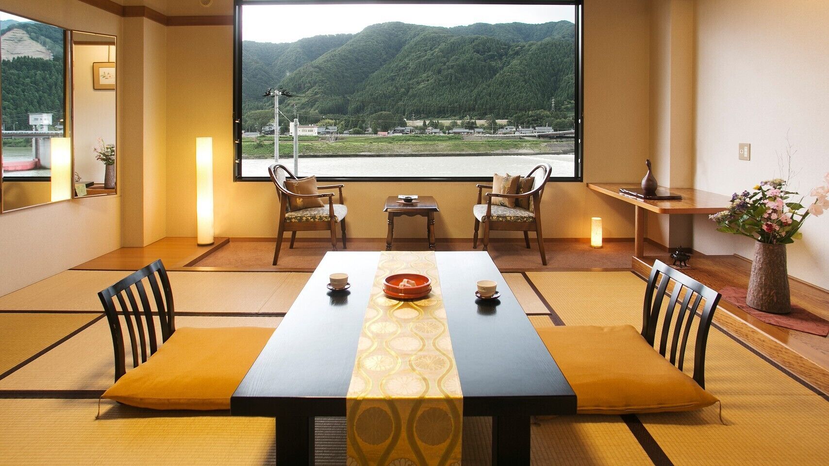 A traditional Japanese room with tatami flooring, a low table with a bowl, chairs, and a view of a mountain and river.