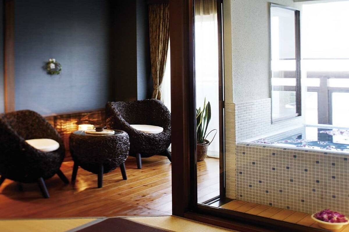Une pièce avec un plancher en bois, deux fauteuils en osier foncé, une petite table, une plante en pot et, par une porte vitrée, une vue sur un jacuzzi.