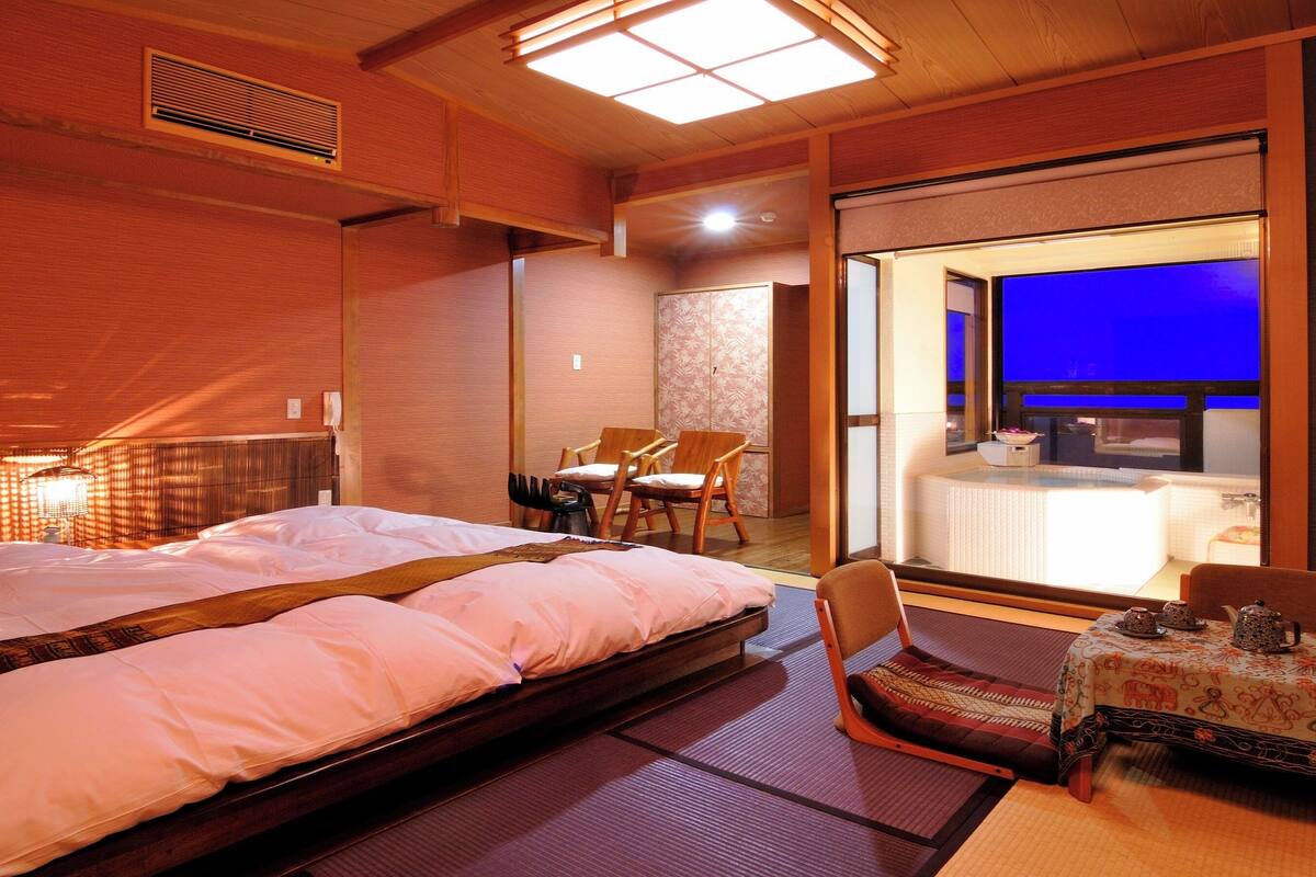 Une chambre d’hôtel avec un grand lit, un coin salon avec des fauteuils, une petite table avec un service à thé, et une vue sur une pièce éclairée de lumière bleue, visible à travers une porte en verre.