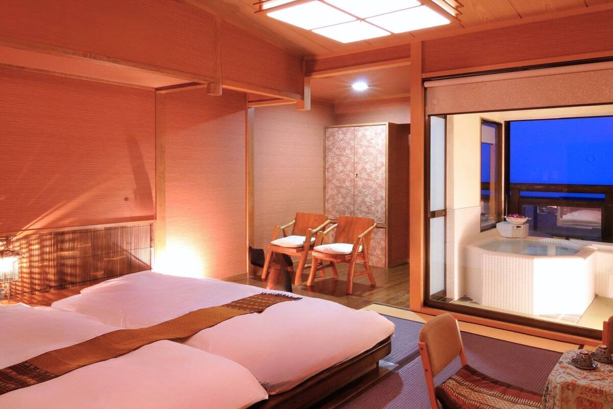 Une chambre d’hôtel dotée d’un grand lit, d’un coin salon avec des fauteuils, d’un jacuzzi fermé par des parois de verre et offrant une vue sur le ciel nocturne.