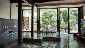 Ein traditionelles japanisches Onsen mit Steinboden, Holzbalkendecke und Blick auf einen üppigen Garten durch große Glasüren.