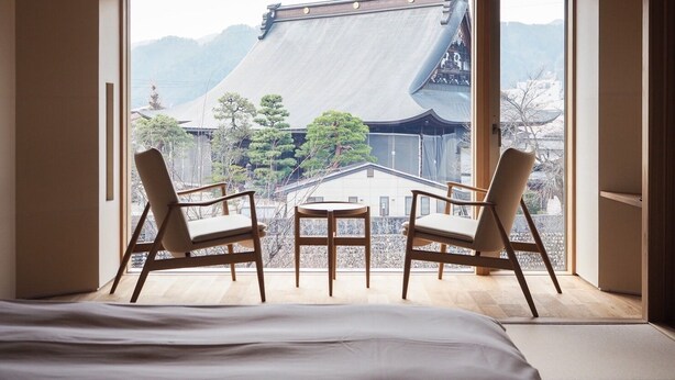 Ein Zimmer mit einem Bett, zwei Stühlen, einem kleinen Tisch und Blick auf traditionelle Architektur.