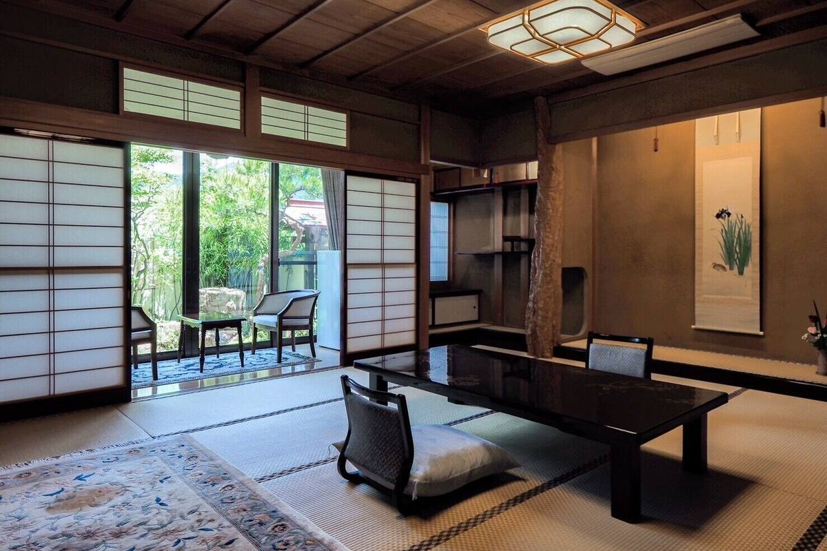 Une pièce de style japonais traditionnel, avec un sol recouvert de tatamis, des portes coulissantes en papier de riz (shoji), une table basse avec des coussins et une vue sur la verdure à l’extérieur.