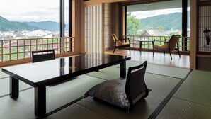 Ein traditioneller japanischer Raum mit Tatami-Bodenbelag, einem niedrigen Tisch und Stühlen. Große Fenster bieten Blick auf eine gebirgige Landschaft.