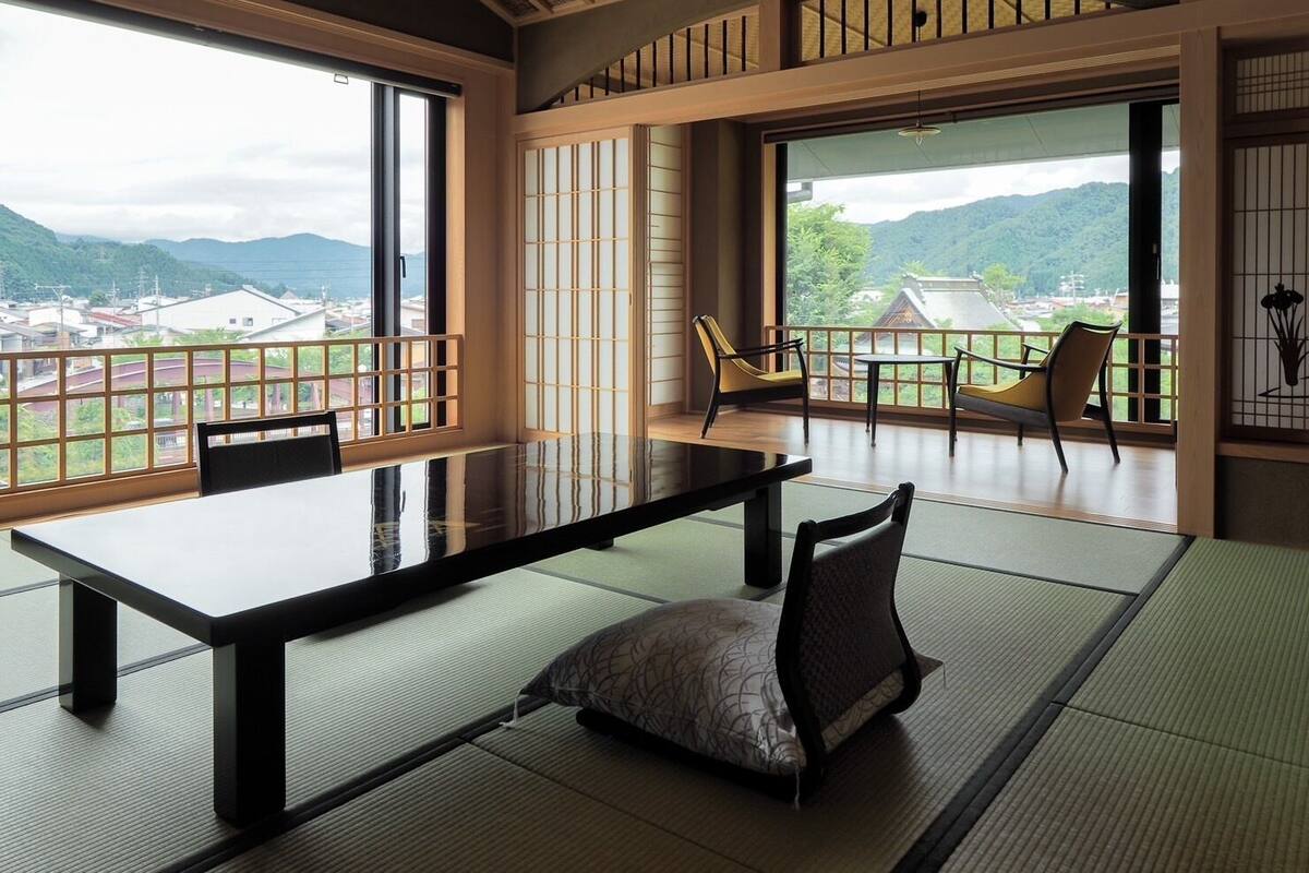 Une pièce de style japonais traditionnel, avec un sol recouvert de tatamis, une table basse et des chaises. De grandes fenêtres offrent une vue sur un paysage montagneux.