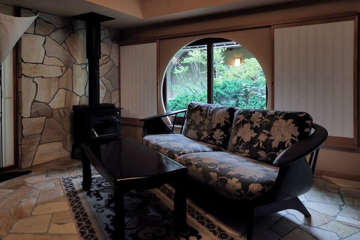 Un salon chaleureux avec une cheminée en pierre, un canapé agrémenté de coussins à motifs floraux et une grande fenêtre donnant sur un paysage verdoyant.