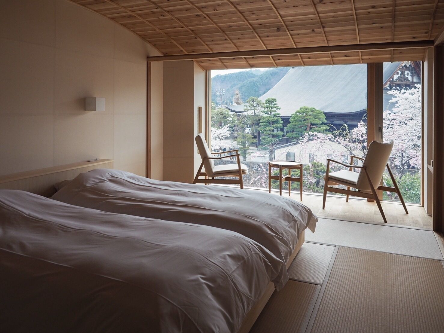 A bedroom with a large bed, two chairs, and a view of cherry blossoms through a sliding door.