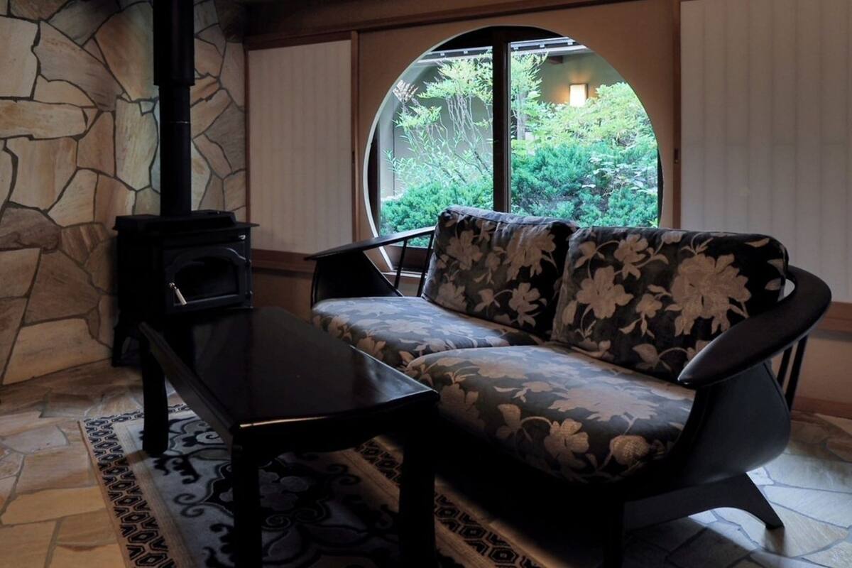 Une cheminée en pierre, un canapé recouvert d’un tissu à motifs floraux et une vue sur la verdure par une grande fenêtre.