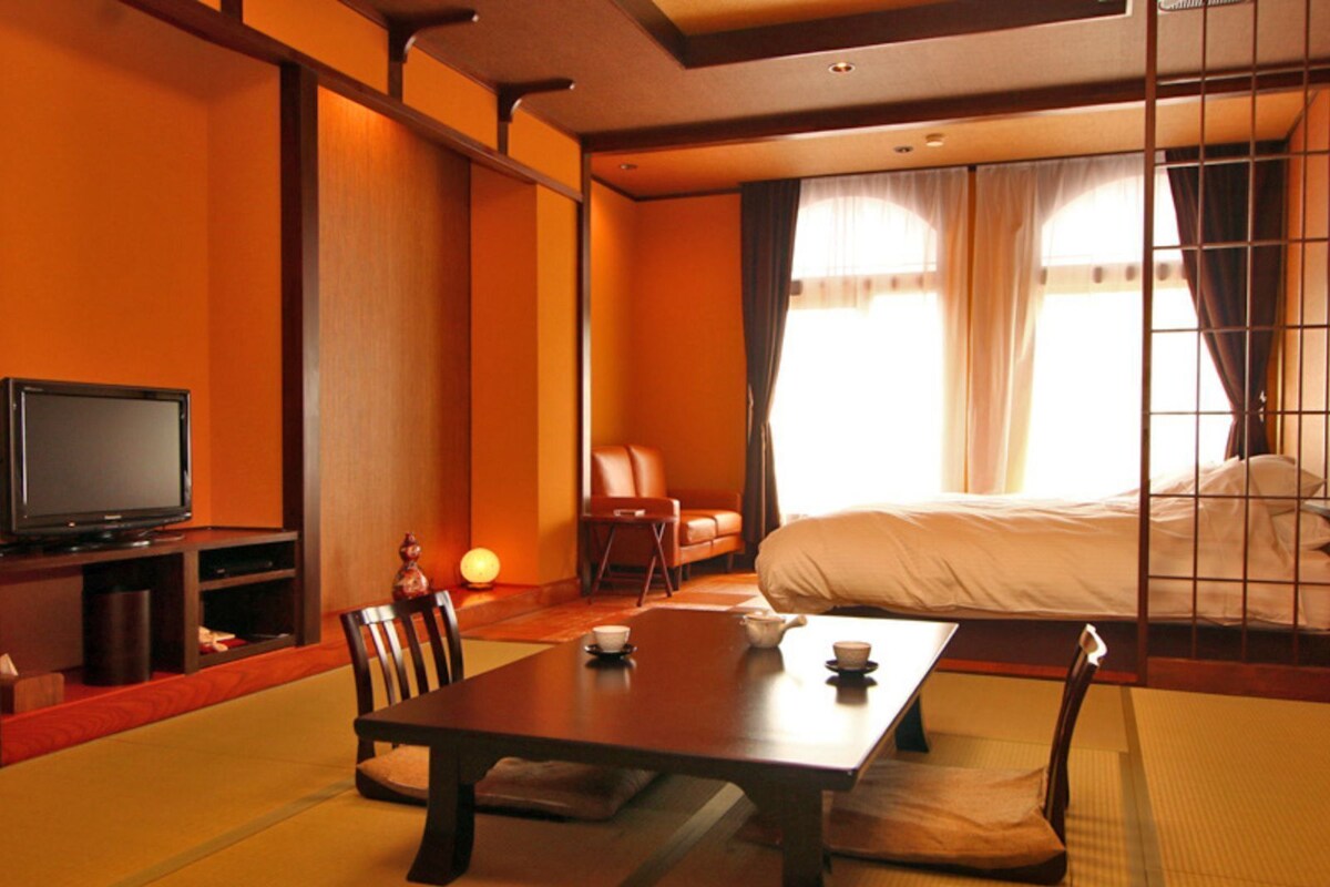 A traditional Japanese-style hotel room with tatami flooring, a low table, a television, a bed, and a sliding door.