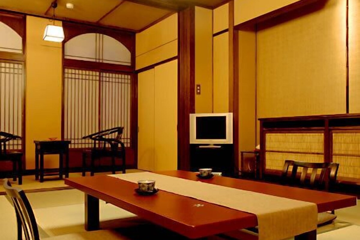 A traditional Japanese room with tatami flooring, a low table, chairs, and a television.