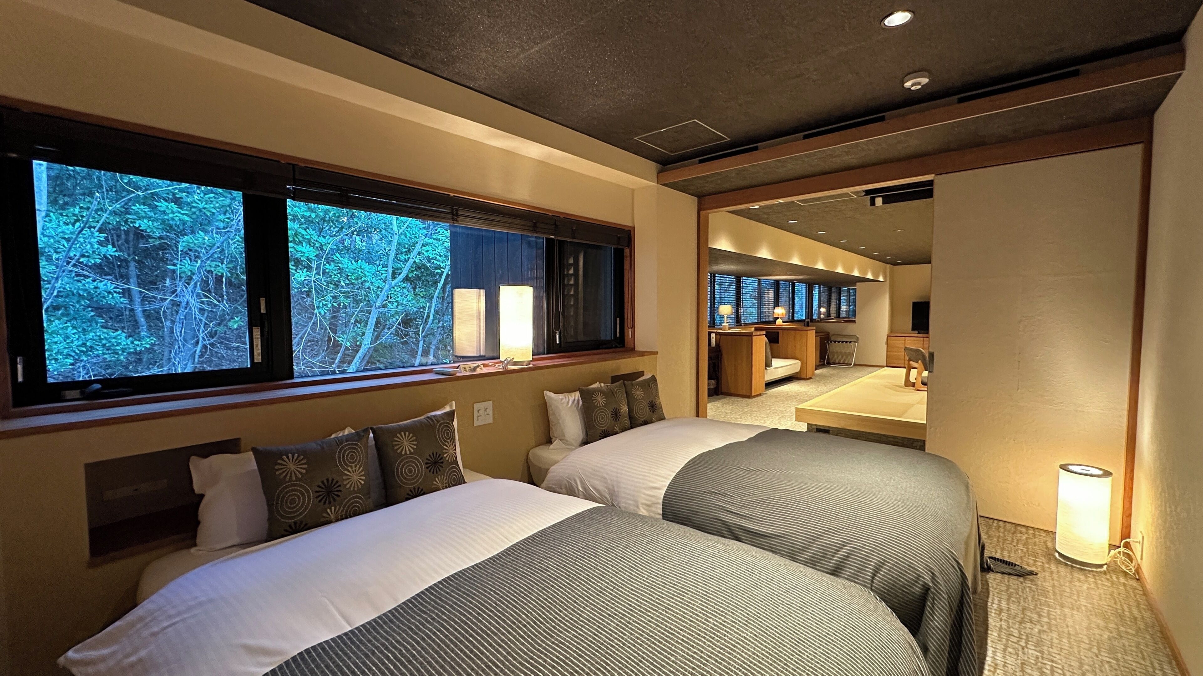 A hotel room with two beds, a large window with a view of trees, a desk with a chair, and a small sitting area.