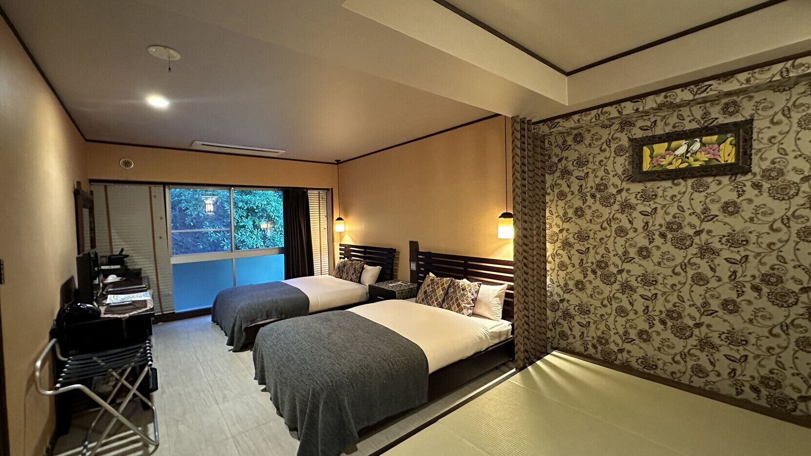 A hotel room with two beds, a desk, and a view of greenery outside.