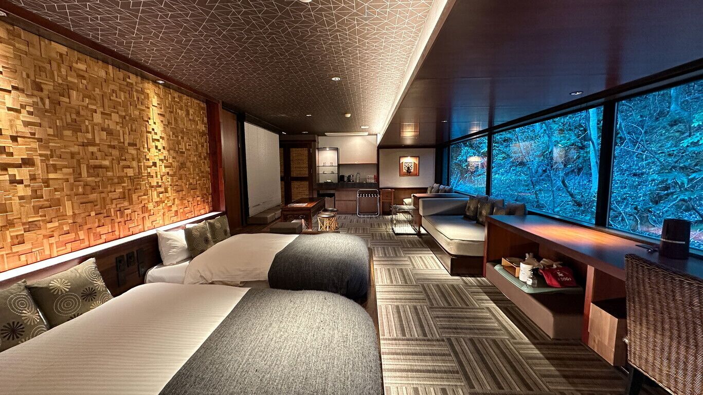 A hotel room with a large window, two beds, a seating area, and a view of a forest.