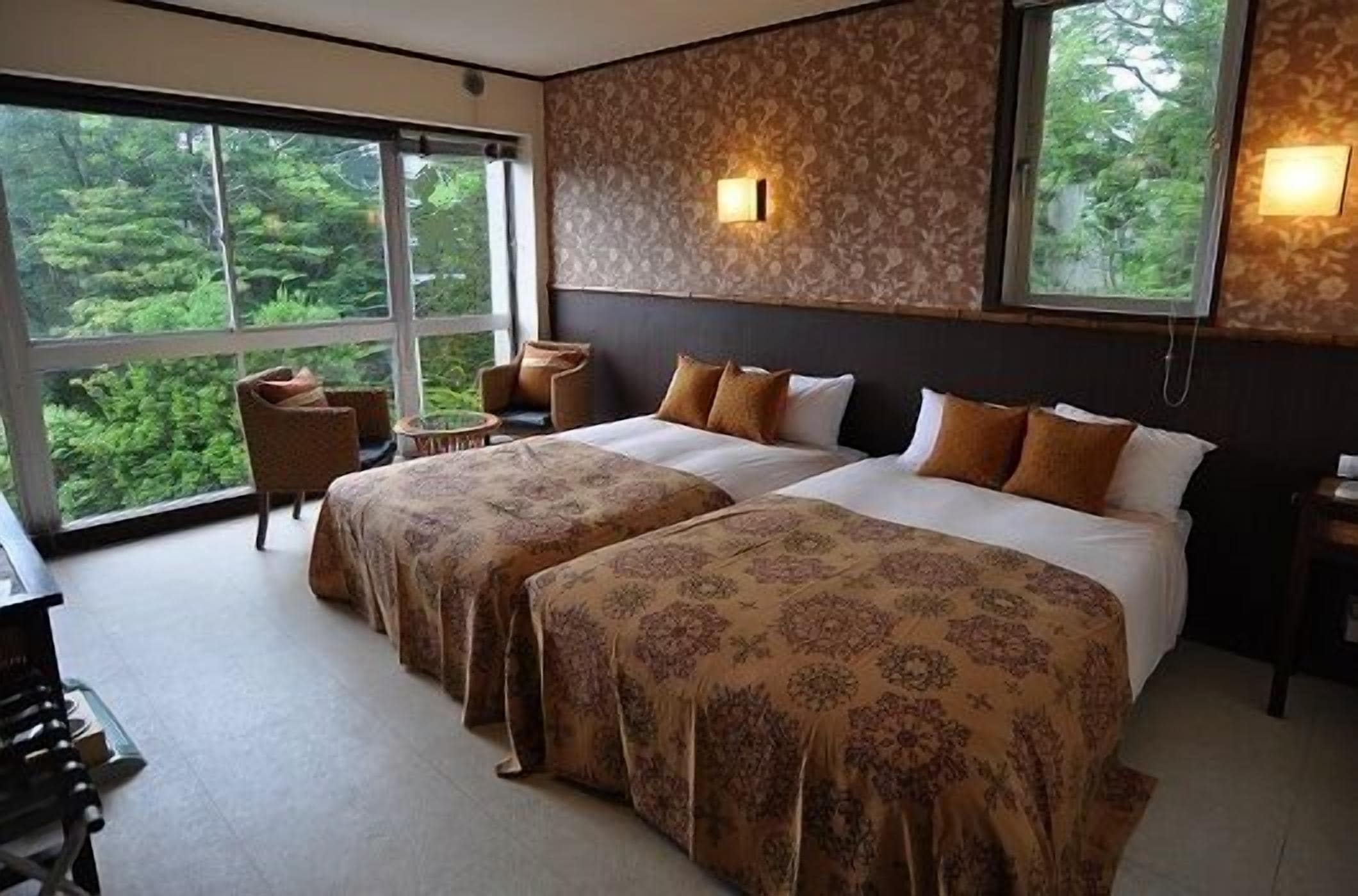 A hotel room with two beds, a sitting area with a chair, a large window with a view of greenery, and a wall-mounted light fixture.