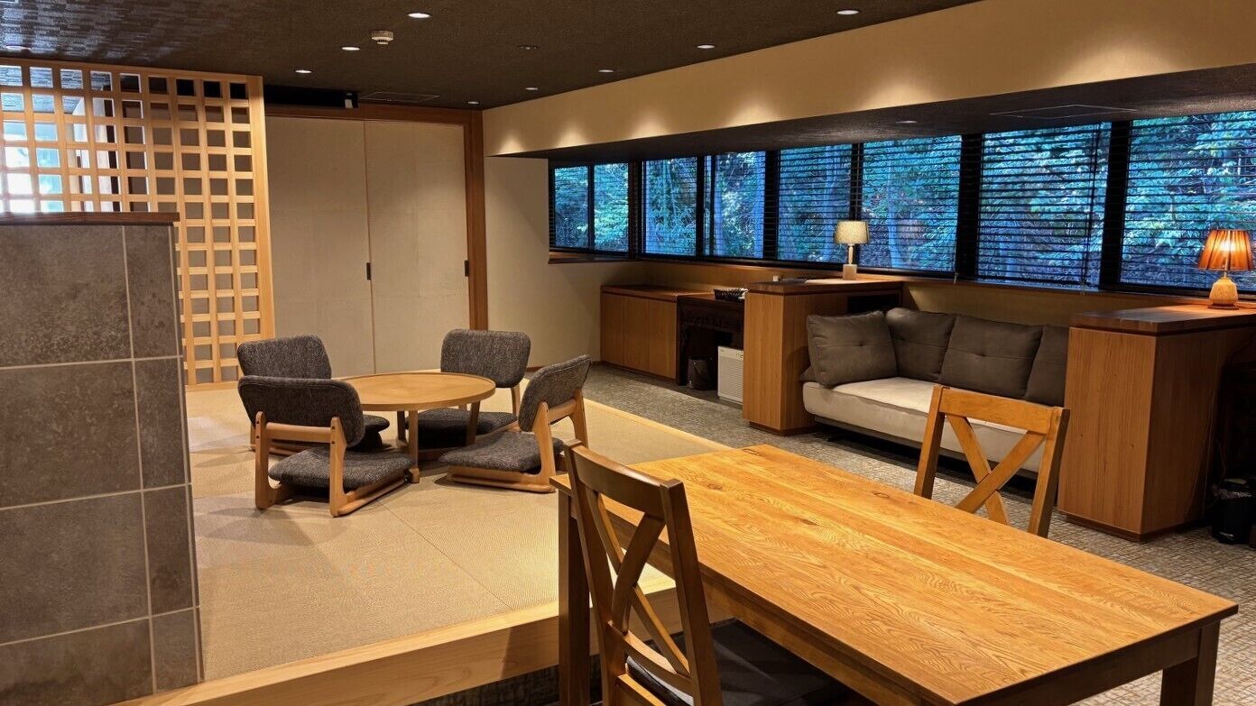 A modern interior with a wooden dining table, grey upholstered chairs, a sofa, and a large window with a view of trees.