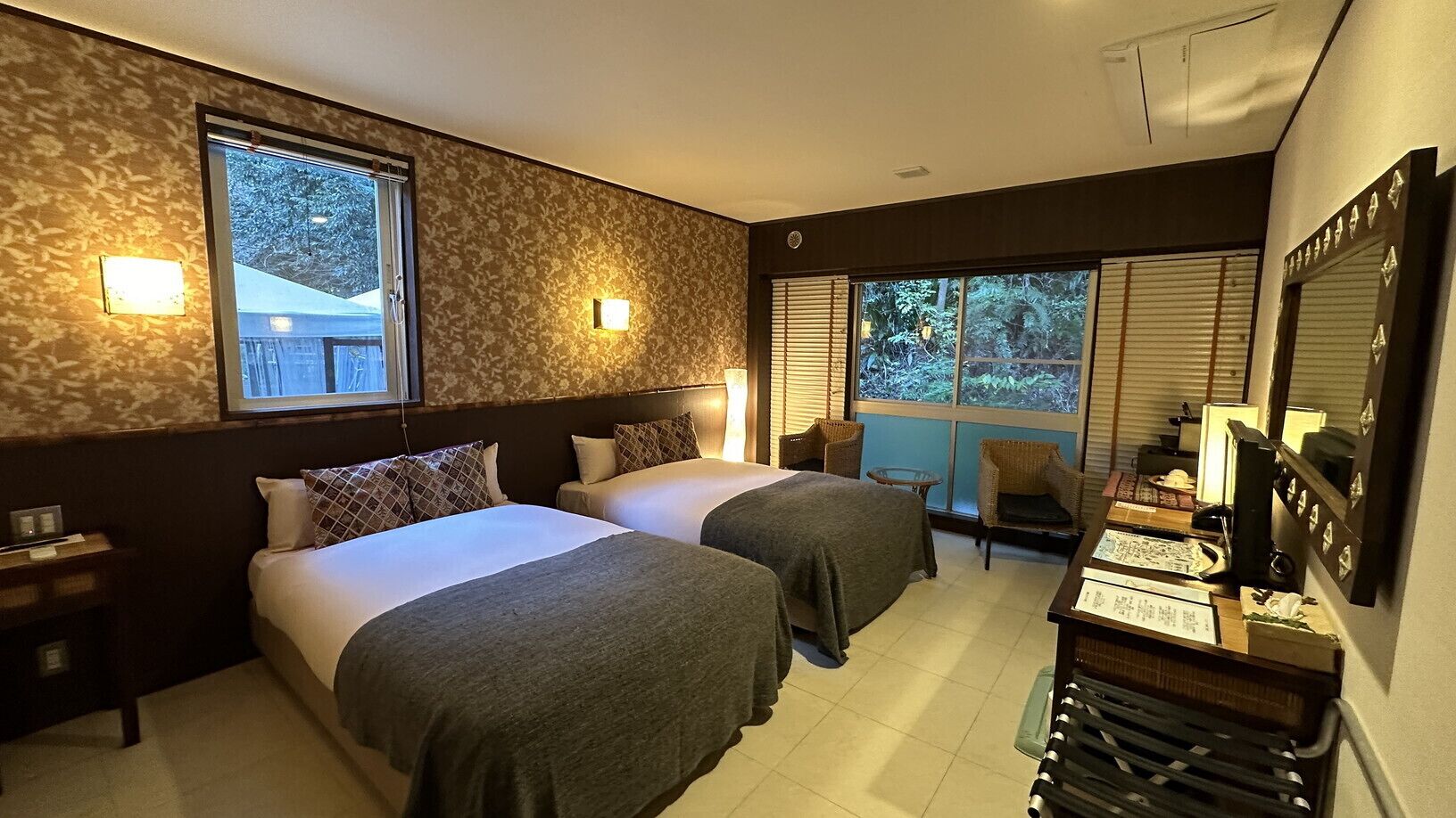 A hotel room with two beds, a desk with a computer, and a large window with blinds.