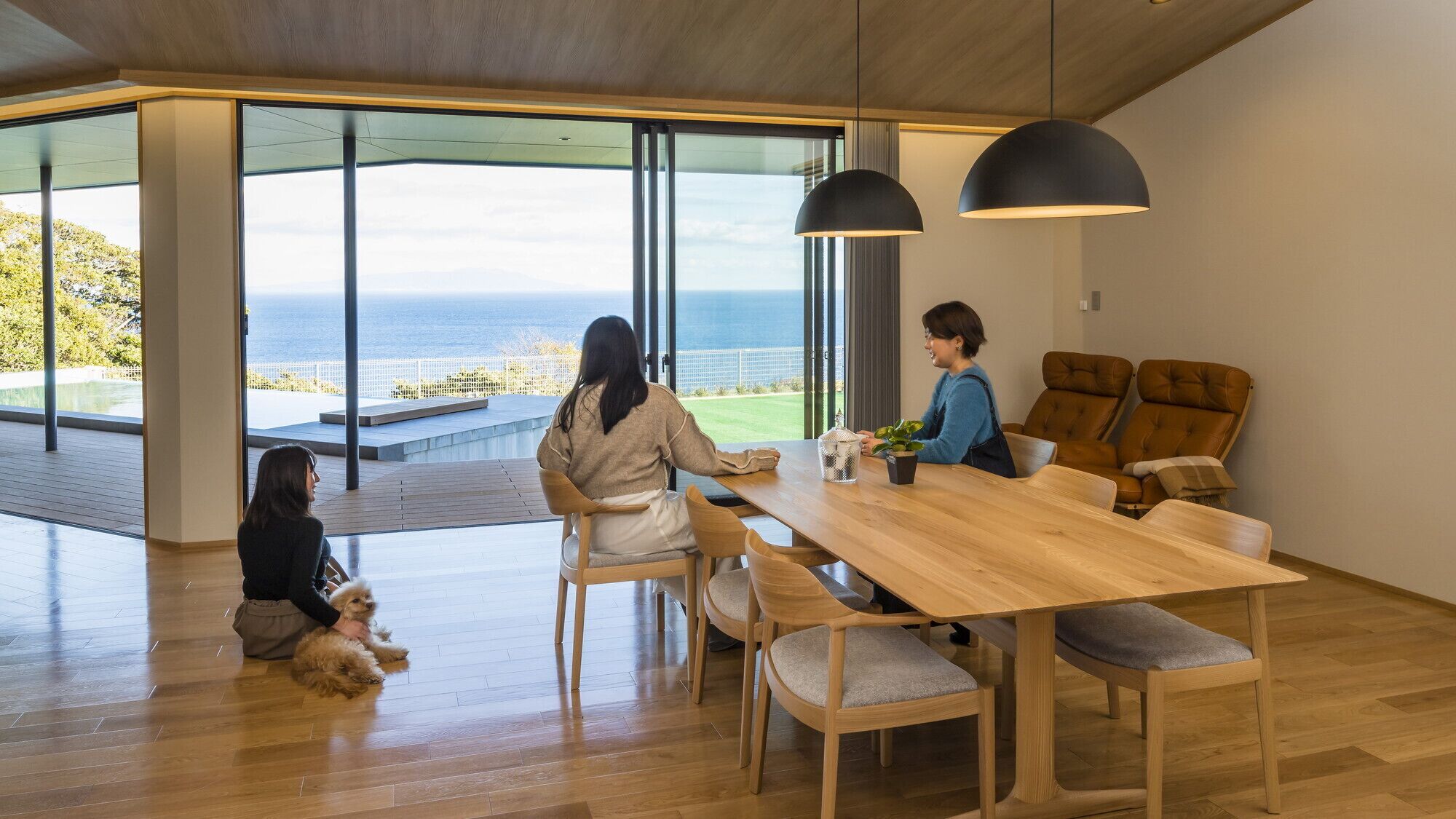A modern interior with a wooden dining table, two chairs, a sofa, and a large window offering a view of the sea.