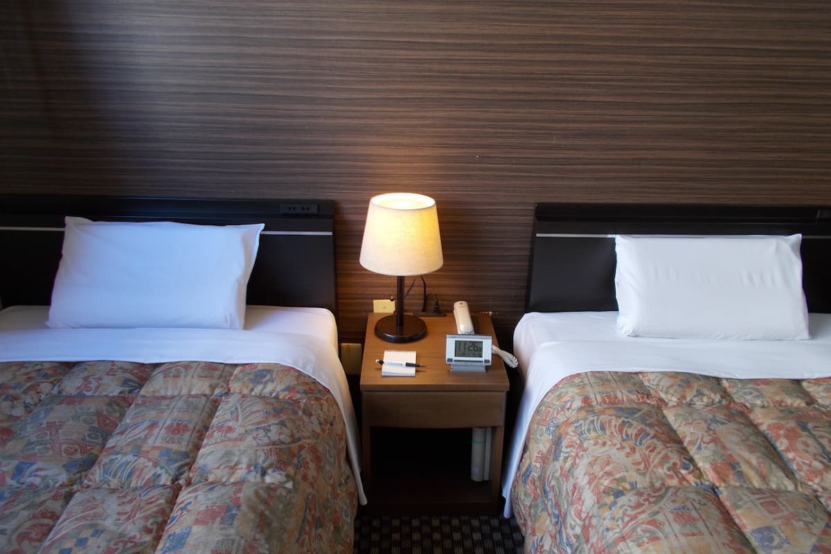 Ein Hotelzimmer mit zwei Betten, jeweils mit einem gemusterten Überwurf, einer Nachttischlampe und einer digitalen Uhr auf dem Nachttisch.