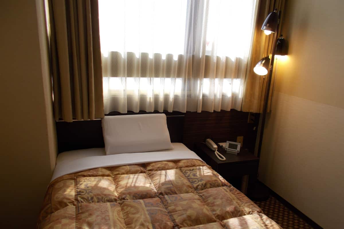 Ein Hotelzimmer mit einem Bett, einem Nachttisch mit Telefon und digitaler Uhr sowie einem Fenster mit Vorhängen.