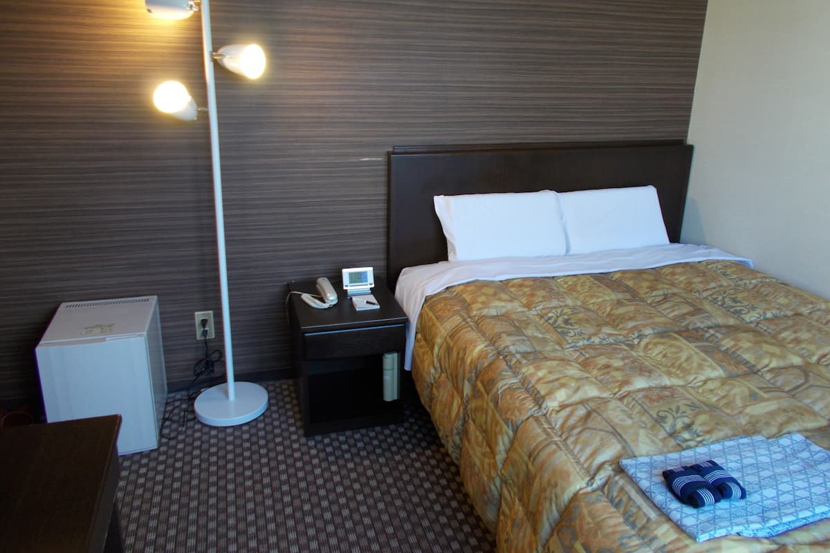 Ein Hotelzimmer mit einem Bett, einem Nachttisch mit Telefon und digitaler Uhr, einem Schreibtisch mit Computer und einer Stehlampe.