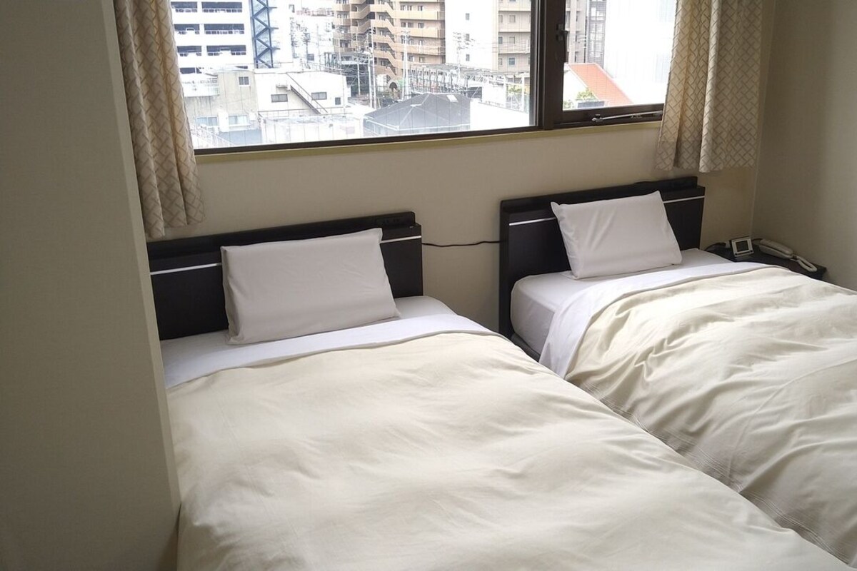 Habitación de hotel con dos camas, una ventana con cortinas, una mesita de noche con teléfono y vistas a edificios.