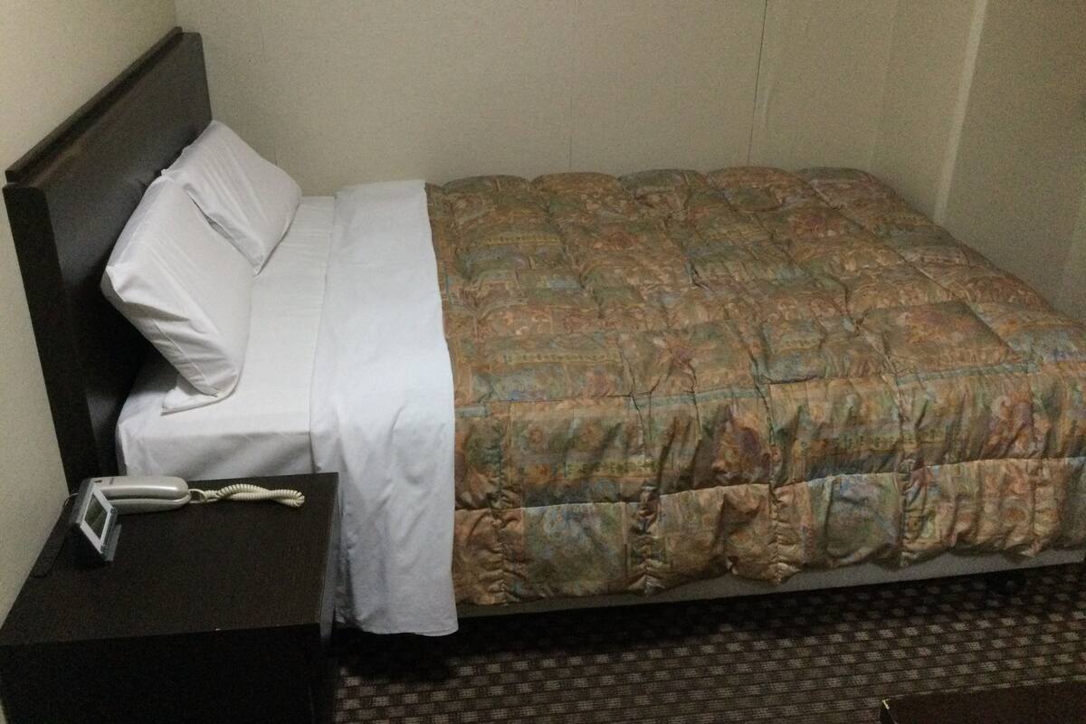 Ein Einzelbett mit einem gemusterten Bettbezug, einem weißen Kopfkissen und einem hölzernen Kopfteil. Ein kleiner Nachttisch mit Telefon und Lampe steht daneben.