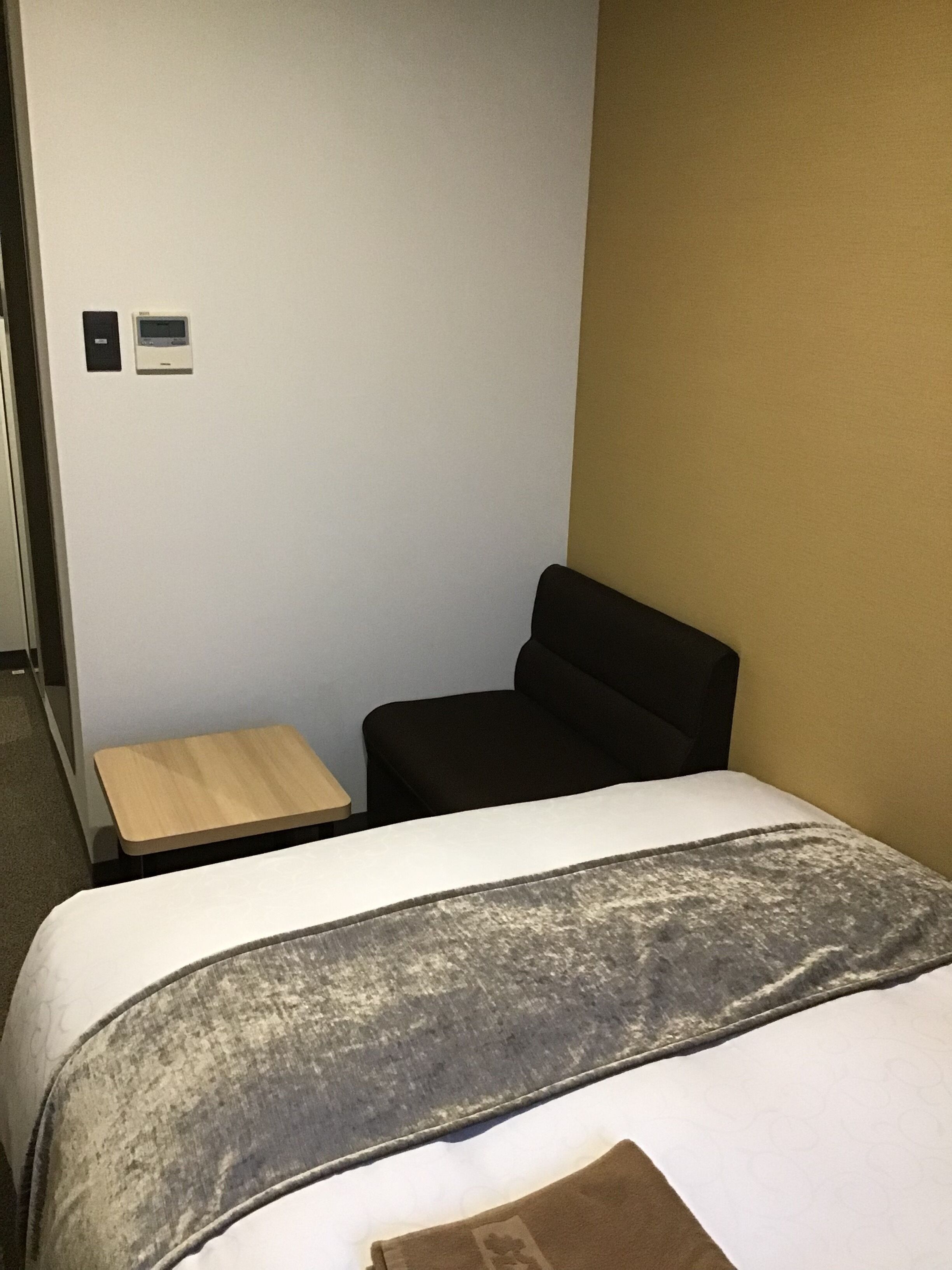 Una habitación de hotel con una cama, una mesita de madera, una silla negra y un panel de control en la pared.