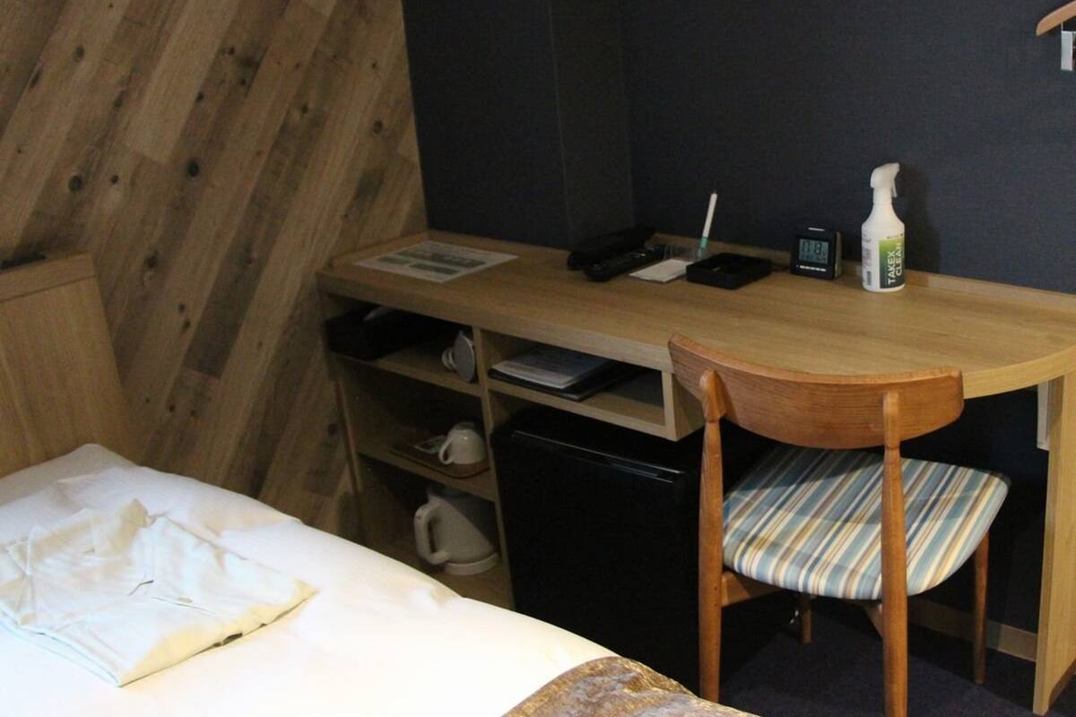 A hotel room with a bed, a desk with a chair, a small shelf, and a wall-mounted telephone.