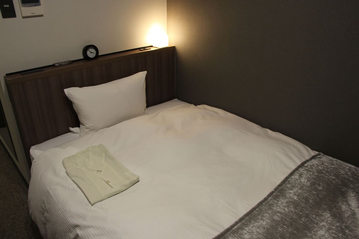 A neatly made bed with a pillow and a folded shirt on it, a clock on the headboard, and a wall-mounted light.