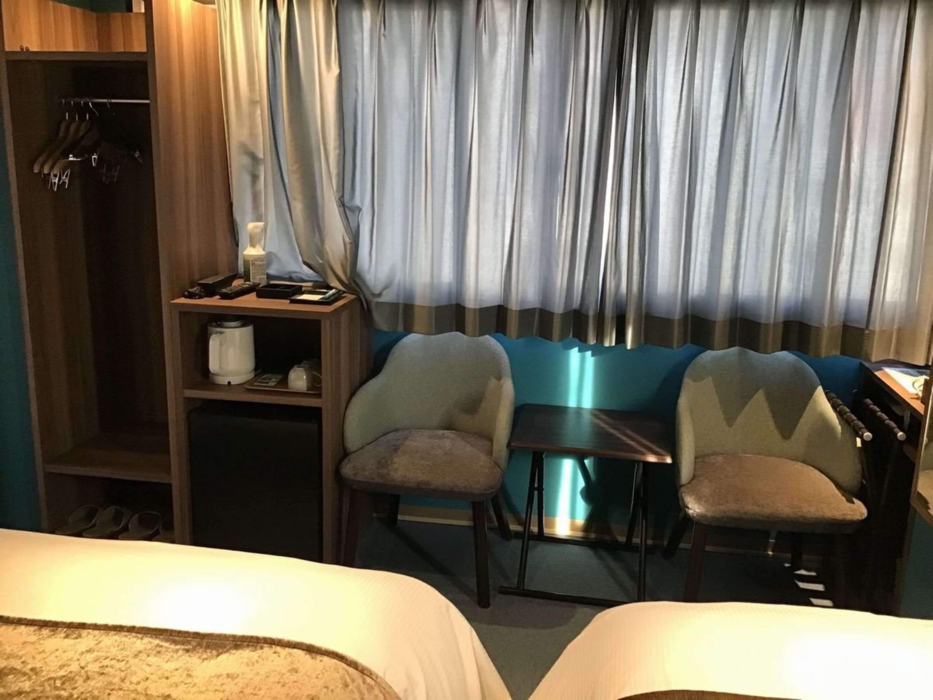 Hotellihuone, jossa on kaksi sänkyä, pieni pöytä, kaksi tuolia, työpöytä, lamppu ja vaatekaappi.