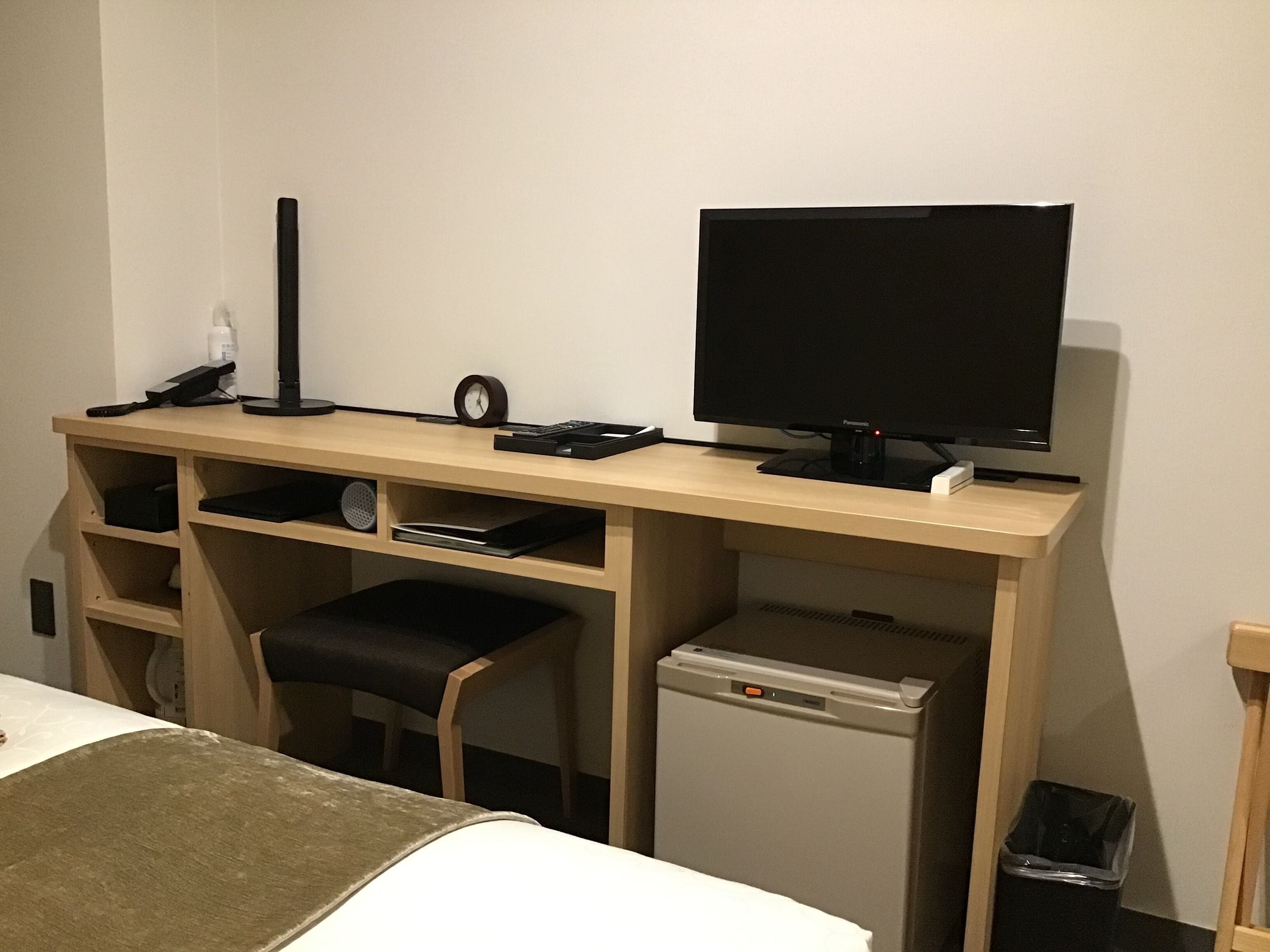 A hotel room with a wooden desk, a television, a small refrigerator, a trash bin, and a bed with a brown blanket.
