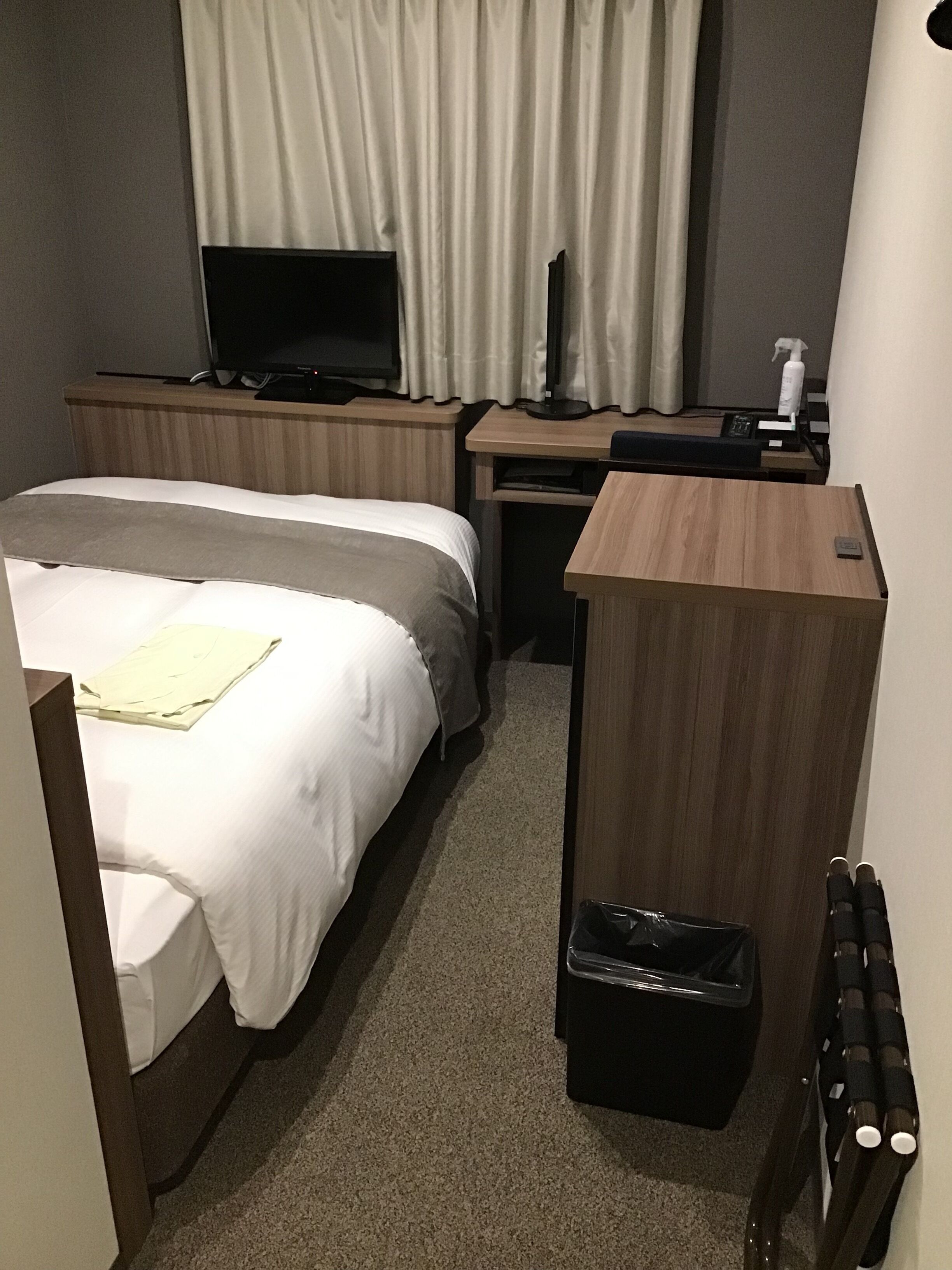 A hotel room with a bed, a desk with a computer, a TV, and a trash bin.