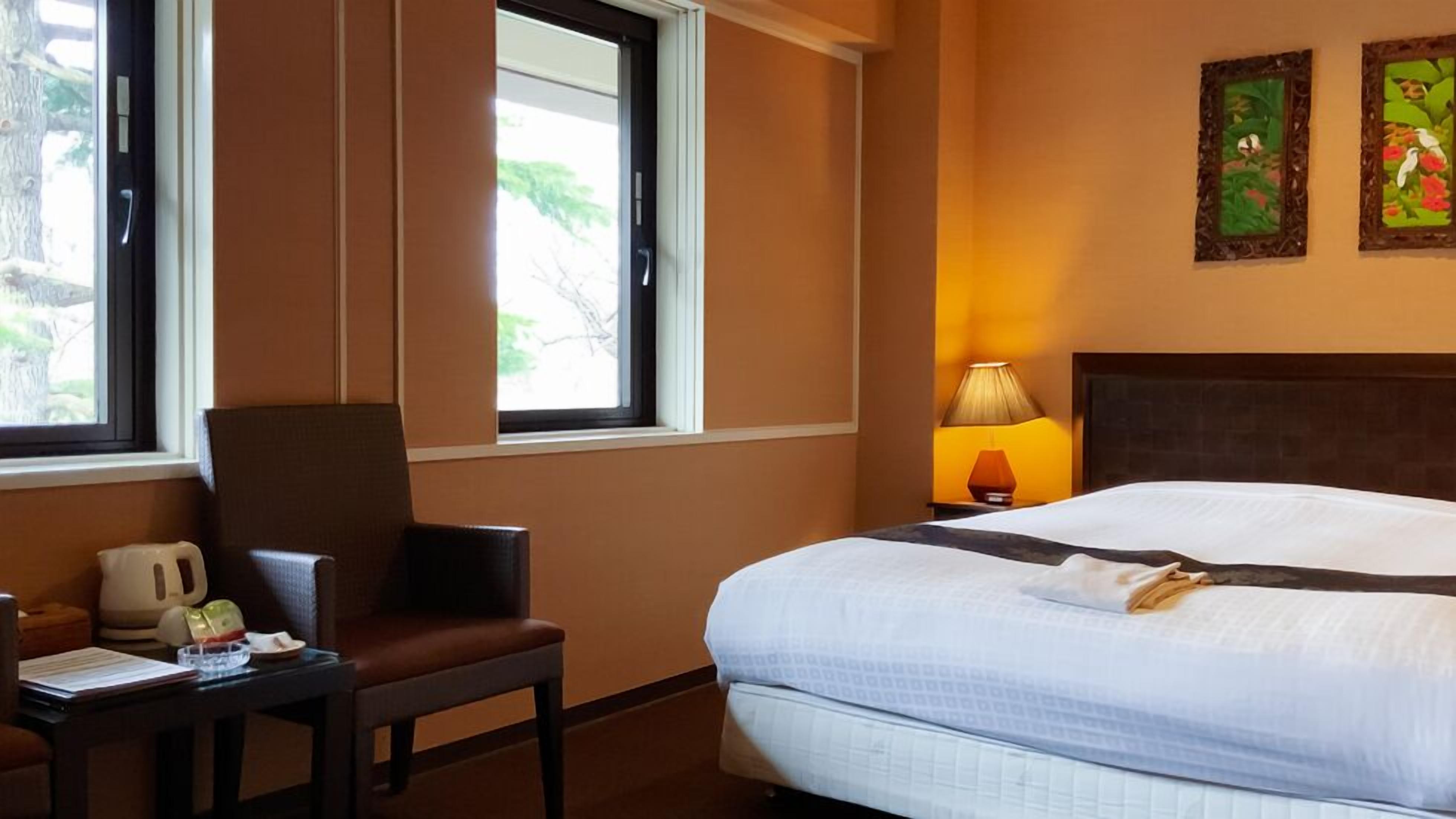 Ein Hotelzimmer mit einem Bett, einem Sessel, einem Schreibtisch mit Wasserkocher, einer Lampe und zwei gerahmten Bildern an der Wand.