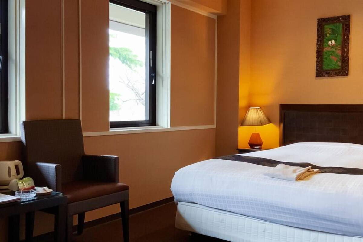 A hotel room with a bed, a chair, a desk with a kettle, a lamp, and two framed pictures on the wall.