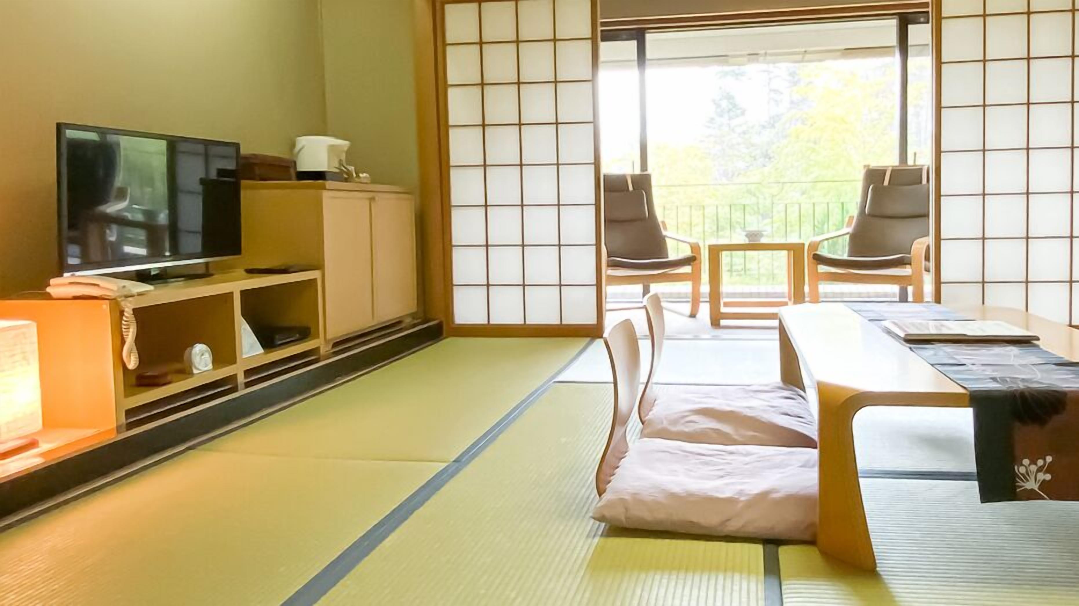 Ein traditioneller japanischer Raum mit Tatami-Böden, einem Fernseher, einem Schrank und einer Schiebetür, die auf einen Balkon mit Stühlen führt.